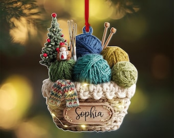 May include: A Christmas ornament shaped like a basket filled with yarn balls in shades of blue, green, and yellow. The ornament includes knitting needles, a miniature Christmas tree, and a small snowman. The name "Sophia" is displayed on a wooden sign.