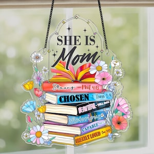 May include: Clear acrylic hanging sign with the words "SHE IS Mom" and a stack of books. The books are decorated with floral accents and inspirational words like "Strong" and "Victorious". The sign is suspended by a black chain.