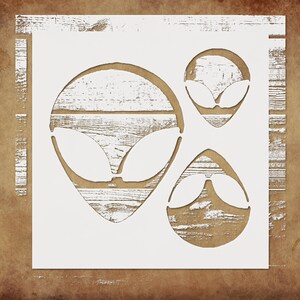 May include: White stencil featuring three alien head designs. The stencil is on a wooden background with a brown and tan color scheme. The alien heads have large oval eyes and a small mouth.