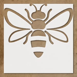 May include: A white stencil featuring a bee design. The bee is cut out of the white material, revealing the brown background. The bee has detailed wings, antennae, and body segments, perfect for crafts and DIY projects.