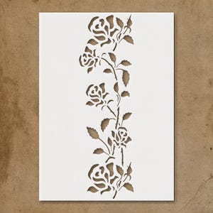 May include: A white stencil featuring a vertical design of roses and leaves. The roses are in various stages of bloom, with detailed petals and stems. The stencil is cut out, creating a negative space design.