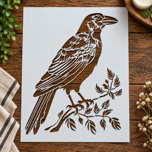 May include: A white rectangular stencil featuring a detailed laser-cut design of a crow perched on a leafy branch. The bird is depicted in profile facing right, with intricate feather patterns and foliage elements visible through the cutouts.