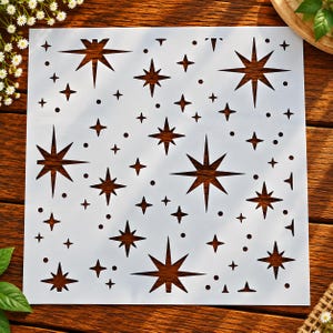 May include: A square white stencil featuring a repeating pattern of various sized stars and small circular dots. The design includes multiple eight-pointed star shapes and smaller four-pointed star shapes cut out from the flat material.