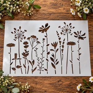 May include: A rectangular white stencil featuring a laser-cut design of various wildflowers and tall grasses. The pattern includes multiple stems, delicate petals, and seed heads arranged in a horizontal row across the flat surface.
