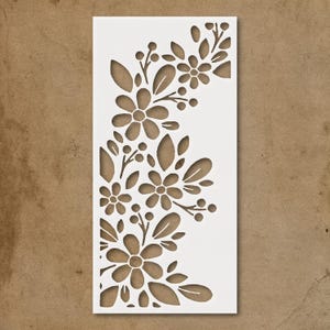 May include: A white stencil featuring a floral design with cut-out flowers, leaves, and berries. The design is arranged in a corner pattern, ideal for crafts and decorative projects. The stencil is rectangular.