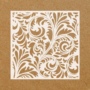 May include: A square, white stencil featuring a detailed, swirling floral design. The intricate pattern includes stylized leaves and curls, set against a textured, brown background. The design is symmetrical and elegant.