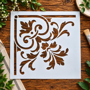 May include: A square white stencil featuring a cutout corner design with intricate acanthus leaf motifs and scrolling flourishes. The stencil has a clean-cut border and a symmetrical pattern that radiates from the corner, creating a decorative template.