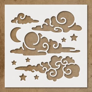 May include: A white stencil featuring a celestial design with clouds, stars, and a crescent moon. The clouds have swirling details, and the stars are five-pointed. The stencil is on a brown background, creating a contrast between the white design and the brown surface.