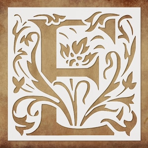 May include: A square stencil featuring the letter "E" in a decorative, ornate style. The letter is surrounded by floral and leaf designs, cut out from a white square. The background is a textured, brown surface.