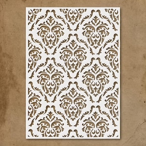 May include: A white stencil with a repeating damask pattern. The design features ornate, symmetrical floral and scrollwork motifs. The stencil is rectangular and appears to be made of a durable, flexible material, suitable for various craft projects.