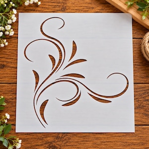 May include: A square, semi-transparent stencil sheet featuring a die-cut design of a flowing, organic corner flourish. The pattern consists of elegant, elongated leaf-like shapes and sweeping, curved lines that originate from the bottom left corner.