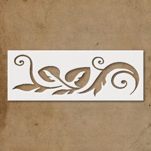 May include: A white stencil featuring a decorative floral design with leaves and swirling elements. The design is cut out, creating a negative space that can be used for painting or other crafts. The stencil is rectangular.