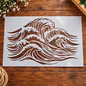 May include: A rectangular stencil featuring a detailed cutout design of multiple crashing ocean waves. The stencil is white and displays intricate, flowing lines that form the crests and foam of the water, creating a repeating wave pattern.