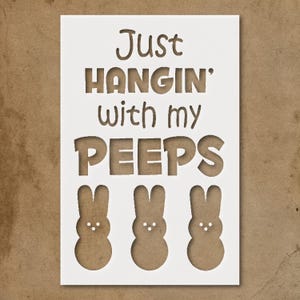 May include: A white stencil with the phrase "Just HANGIN' with my PEEPS" cut out. Below the text are three brown bunny shapes. The stencil is on a brown background.