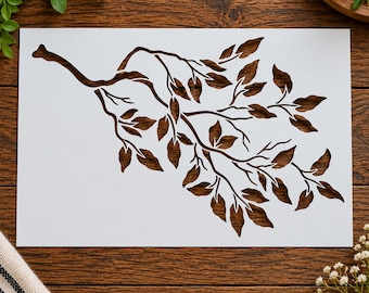 May include: A rectangular white stencil featuring a detailed cutout design of a tree branch with numerous leaves. The branch extends diagonally across the surface, showcasing intricate stems and various leaf shapes in a negative space pattern.