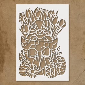 May include: A white paper cut-out design featuring an Easter bunny surrounded by tulips and decorated Easter eggs. The bunny is in a basket with a bow. Daisies and leaves are also included in the design.