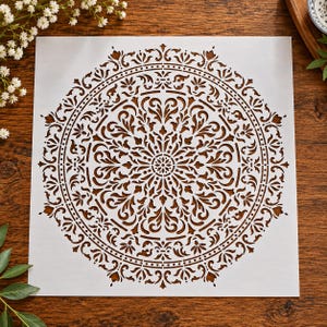 May include: A square white stencil featuring a large, intricate circular mandala design. The pattern consists of symmetrical, repeating floral and leaf motifs radiating from a central point, with cut-out sections creating a detailed lace-like effect.