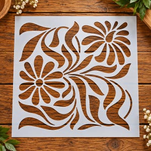 May include: A square, semi-transparent white stencil featuring a cutout design of two large, stylized flowers with elongated, flowing petals and curved, sweeping leaves that fill the entire surface area of the square template.