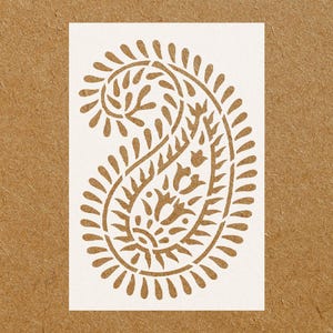 May include: White stencil of a paisley design with intricate details. The design is a traditional Indian pattern with a teardrop shape and floral motifs.