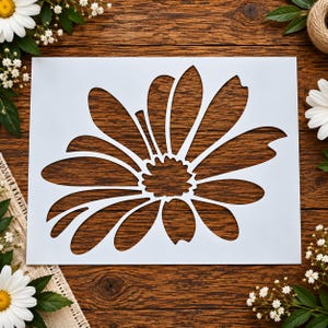 May include: A rectangular white stencil featuring a large, die-cut daisy flower design with a central textured disc and numerous elongated petals radiating outward, creating a clear negative space pattern for painting or crafting applications.