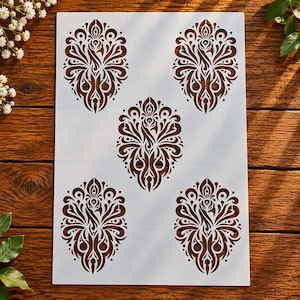 May include: A rectangular white stencil sheet featuring five identical, symmetrical, intricate peacock-inspired damask patterns arranged in a quincunx layout, with cutouts forming the detailed swirling and feathered motifs.