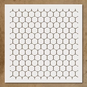 May include: A white stencil featuring a repeating honeycomb pattern. The honeycomb cells are outlined in a dark brown color, creating a geometric design. The stencil is on a light brown surface.