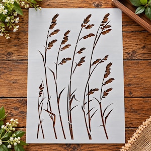 May include: A rectangular white stencil featuring a detailed cutout design of multiple tall grass stalks with slender stems and textured seed heads. The stencil is oriented vertically, showcasing a repeating pattern of wild meadow grasses.