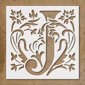 May include: A white stencil featuring the letter "J" in an ornate, floral design. The letter is surrounded by stylized flowers, leaves, and swirling patterns. The stencil is square and set against a brown background.