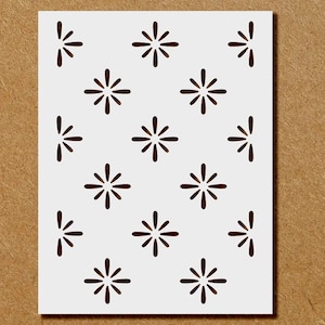 May include: A white stencil with a repeating pattern of dark brown starburst shapes. The stencil is rectangular and sits on a brown background. The starbursts are evenly spaced across the stencil's surface.