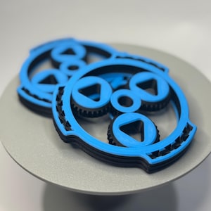 Può includere: Due oggetti stampati in 3D blu e neri con un design circolare, caratterizzati da elementi triangolari e simili a ingranaggi. Gli oggetti sono stratificati e poggiano su una superficie grigia e rotonda. La combinazione di colori è principalmente blu e nero.