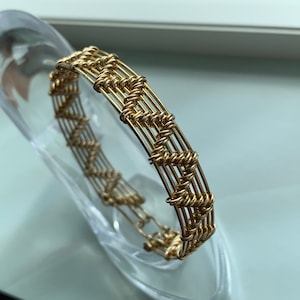 Puede incluir: Un brazalete de alambre dorado con un diseño tejido. El brazalete está hecho de varios hilos de alambre que se tejen juntos para crear un patrón texturizado.
