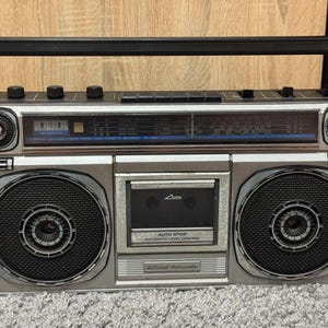May include: A silver and black portable cassette player and radio boombox featuring a top handle, two large circular speakers, a central cassette deck with Auto Stop and Automatic Level Control text, and a radio tuner display with frequency scales.