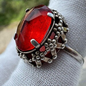 May include: A silver ring featuring a large, oval-shaped, faceted red stone set in a metal basket. The setting is decorated with a repeating pattern of small, raised flower shapes around the perimeter, securing the stone with multiple prongs.