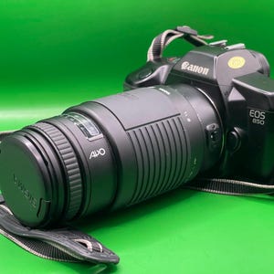 May include: A black Canon EOS 850 camera with a Sigma lens. The lens is capped and has the text "SIGMA" on it. The camera strap is black and white. The background is green.