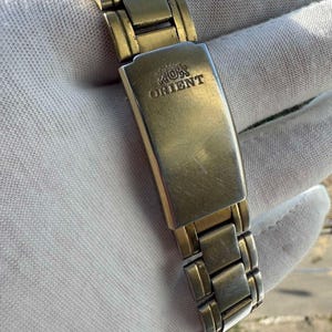May include: Close-up of a gold-toned metal watch band with the word "ORIENT" engraved on the clasp. The watch band is composed of rectangular links and is set against a white background.