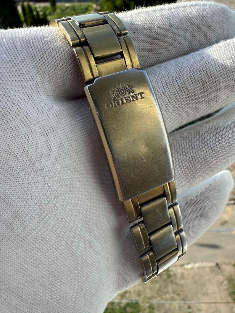 May include: Close-up of a gold-toned metal watch band with the word "ORIENT" engraved on the clasp. The watch band is composed of rectangular links and is set against a white background.