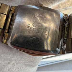May include: Close-up of the back of a vintage Orient watch. The watch has a rectangular stainless steel case and a gold-toned metal band. The watch face has the Orient logo and text that reads "KY PMAA-40 CS WATER RESISTANT 50M STAINLESS STEEL ORIENT WATCH CO., LTD."