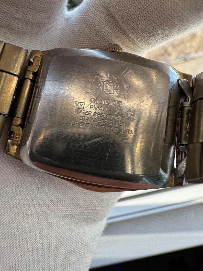 May include: Close-up of the back of a vintage Orient watch. The watch has a rectangular stainless steel case and a gold-toned metal band. The watch face has the Orient logo and text that reads "KY PMAA-40 CS WATER RESISTANT 50M STAINLESS STEEL ORIENT WATCH CO., LTD."
