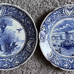 May include: Two round ceramic plates featuring a blue and white transferware pattern. The left plate depicts a hunter with a dog in a marshy landscape. The right plate shows sailboats on water near a shoreline. Both have ornate floral borders.