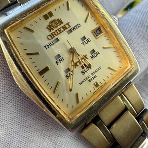 May include: A gold-toned Orient wristwatch with a square face. The watch face displays the days of the week, the date, and the Orient logo. The watch band is also gold-toned.