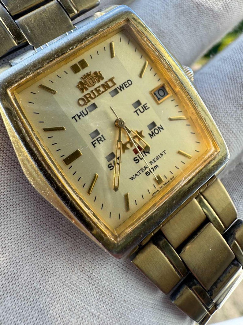 May include: A gold-toned Orient wristwatch with a square face. The watch face displays the days of the week, the date, and the Orient logo. The watch band is also gold-toned.