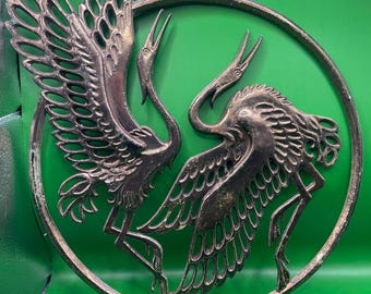 May include: Bronze-colored metal wall art featuring two stylized cranes within a circular frame. The cranes have outstretched wings and long beaks, creating a dynamic and elegant design. The piece has a vintage aesthetic.