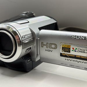 Puede incluir: Videocámara Sony plateada y negra con una lente que indica 4.0 megapíxeles. La cámara tiene una pantalla abatible y una correa. El texto "Full HD 1080" y "ClearVid CMOS sensor" son visibles.
