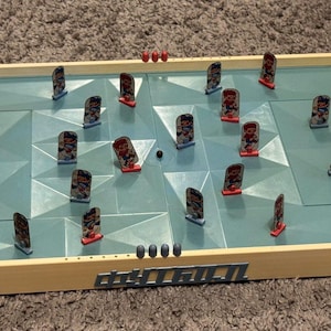 Op de afbeelding: Een ijshockeytafelspel met een crèmekleurig kunststof frame en een lichtblauw, gefacetteerd speeloppervlak. Het bord bevat twee doelen, blauwe bedieningshendels aan elke kant en verschillende kleine speelfiguren op de ijsbaan.