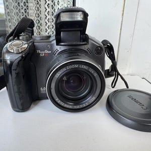 Puede incluir: Una cámara digital Canon PowerShot S3 IS negra con objetivo zoom y tapa de objetivo. La cámara tiene un flash incorporado y una correa. La lente dice "CANON ZOOM LENS 12x IS".