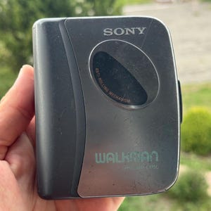 May include: A dark gray Sony Walkman portable cassette player with a metallic finish. The device features the text SONY, WALKMAN, and WM-EX150, along with a circular window labeled ANTI-ROLLING MECHANISM. The surface shows minor surface wear.