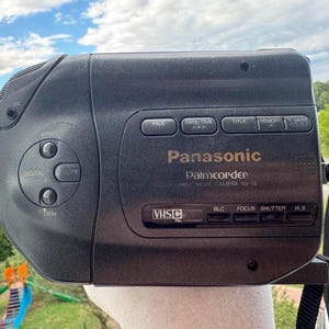 May include: A black Panasonic Palmcorder VHS-C movie camera model NV-S6. The device features a side control panel with buttons for effect, digital, and zoom. The main body displays labels for fade, date/time, title, memory, and mode, with a black strap.