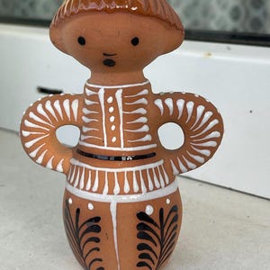 Puede incluir: Figura de terracota con cabeza redondeada y brazos con aberturas circulares. La figura tiene un esmalte marrón en la cabeza y está decorada con detalles pintados en blanco y negro. El cuerpo de la figura es cilíndrico, con un diseño detallado.