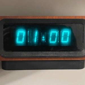 May include: A vintage digital clock displaying 01:00 in bright teal digits. The clock has a rectangular wooden frame with a black border around the display. The base is black, and a white cord extends from the side.