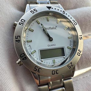 Op de afbeelding: Een zilverkleurig analoog en digitaal polshorloge met een witte wijzerplaat, voorzien van de tekst Westfalia en QUARZ. Het horloge heeft een roestvrijstalen schakelarmband, een draaibare ring met minutenaanduiding en lichtgevende uurindexen.